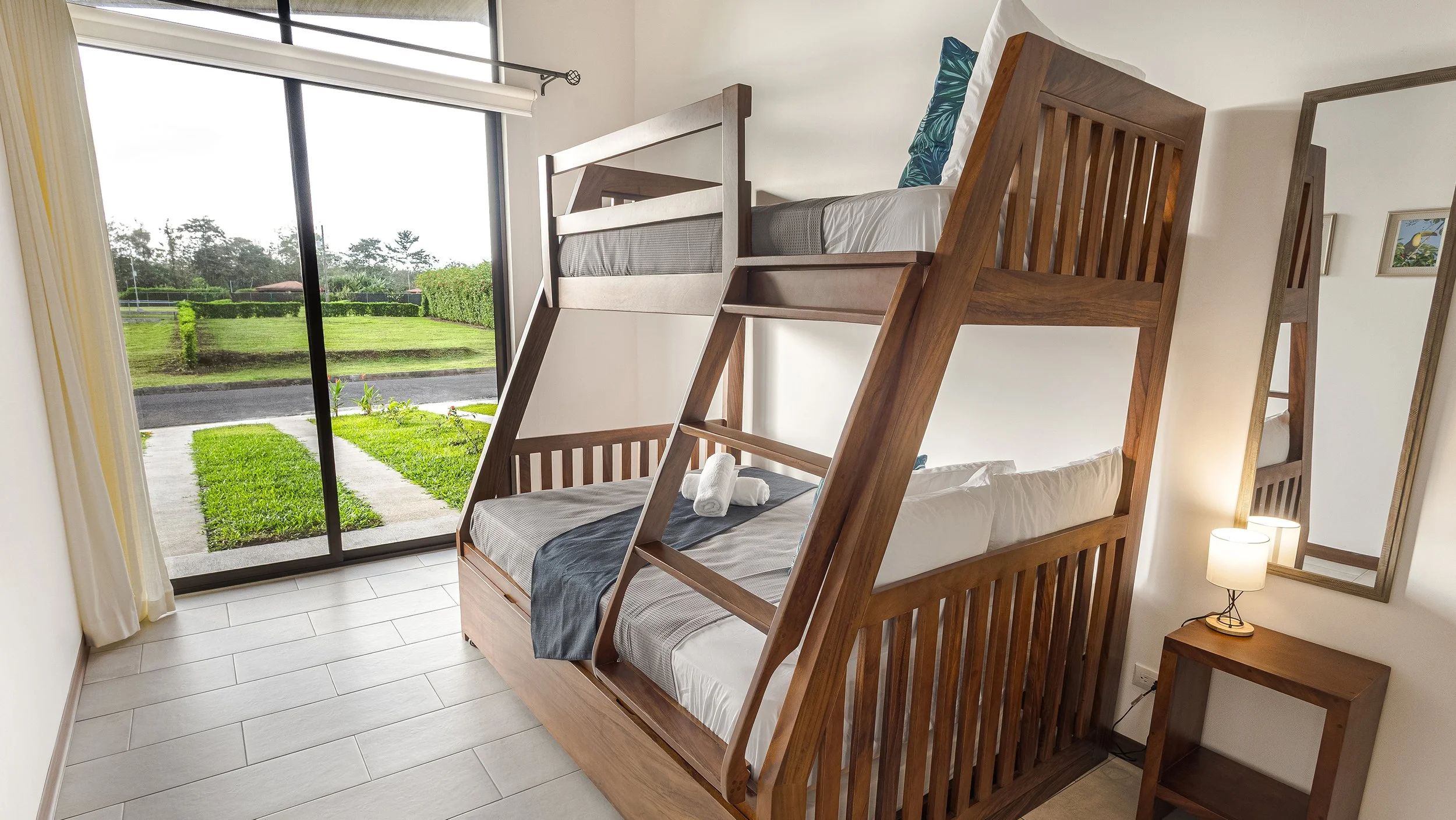 A bedroom with wooden bunk beds near a sliding glass door leading to a green lawn outside. The bunk beds have a roll of towels on the lower bed and a decorative pillow on the upper bed. There is a small nightstand with a lamp and a mirror on the wall