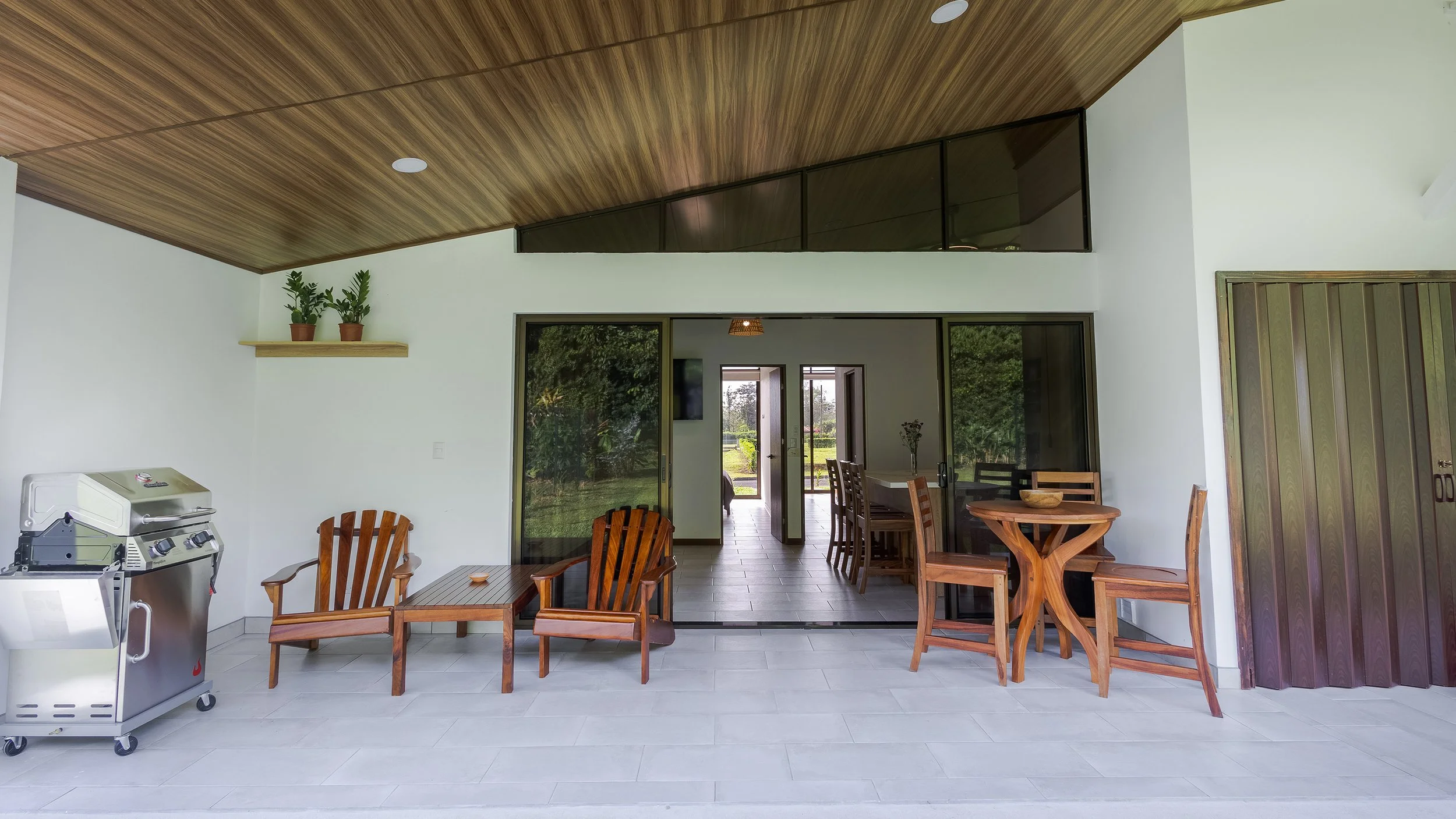Outdoor patio area with wooden dining chairs, a round table, a barbecue grill, and sliding glass doors leading into a house. There are potted plants on a shelf on the white walls, and the ceiling has a wood finish.
