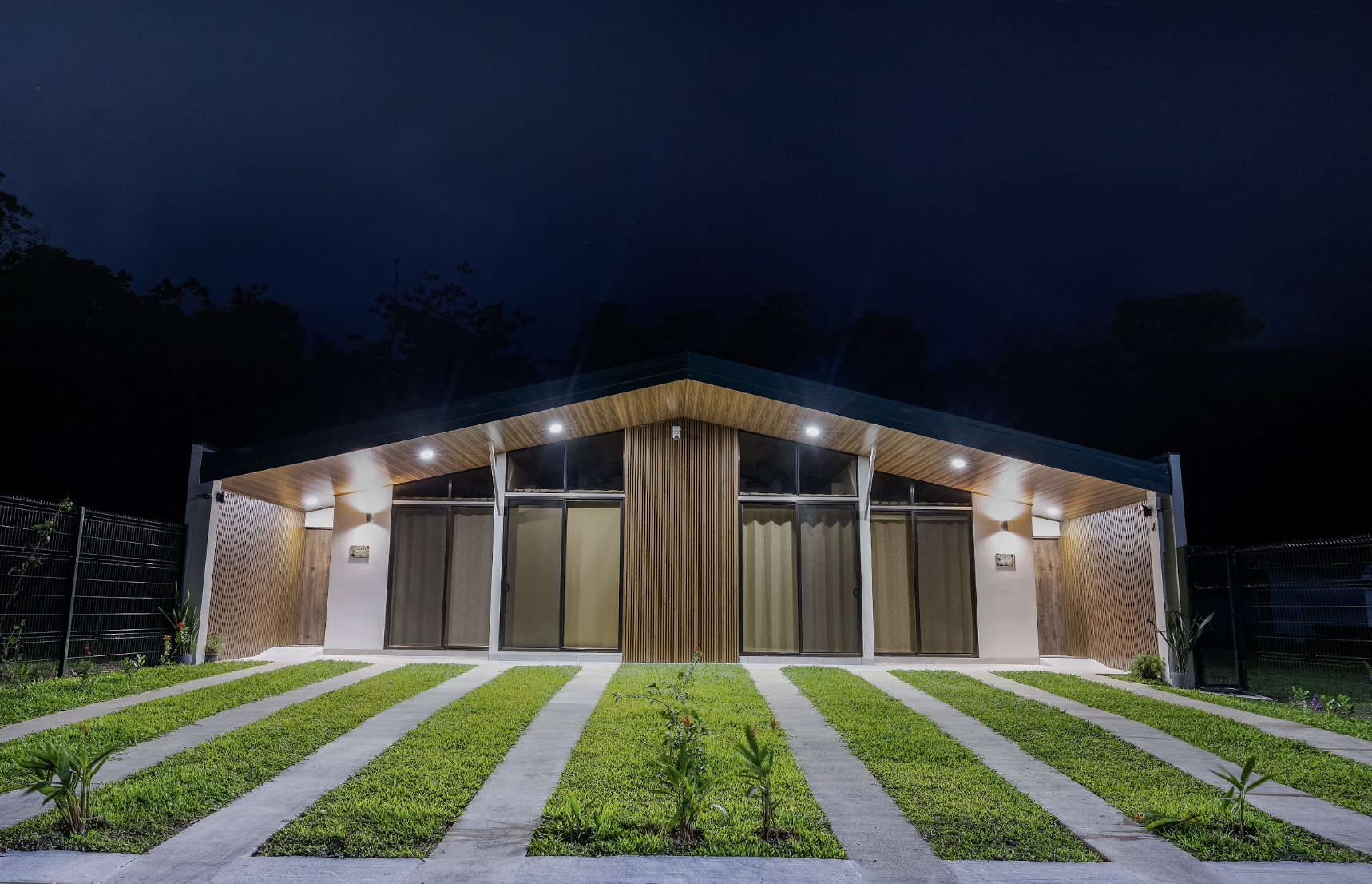 Modern house at night with large glass sliding doors, wooden accents, and exterior lighting, surrounded by a green lawn with stone pathways.