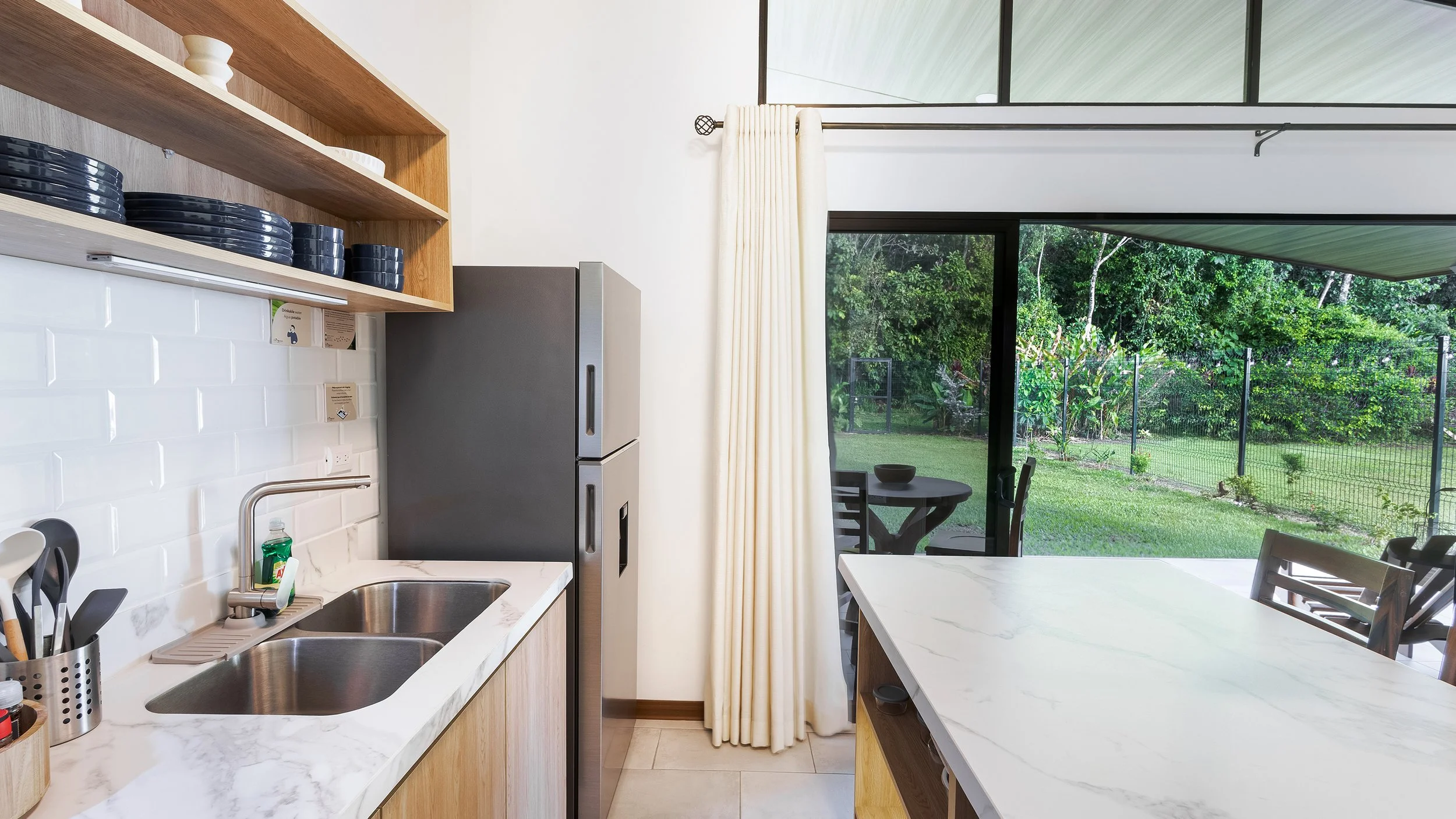 A modern kitchen with a view of a backyard garden through large sliding glass doors, featuring a white marble countertop, wooden shelves, and stainless steel appliances.
