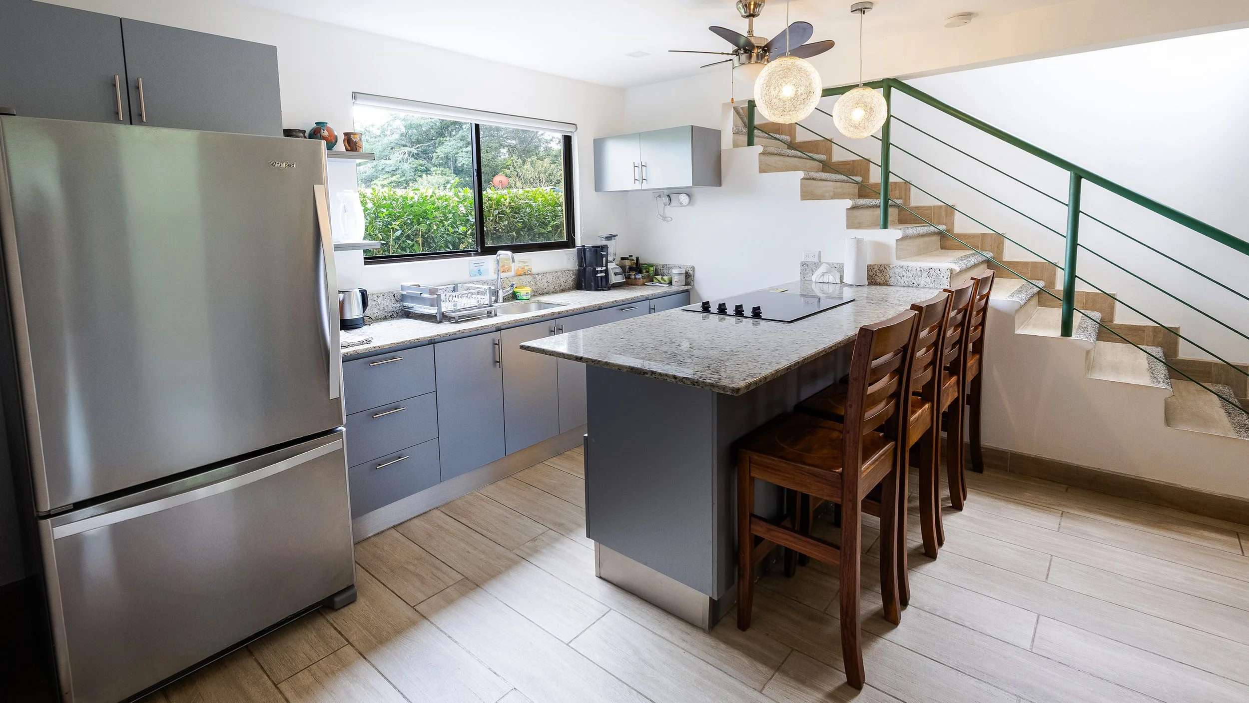 Modern kitchen with gray cabinets, stainless steel refrigerator, granite countertops, and wooden chairs, with a staircase and large window showing greenery outside.