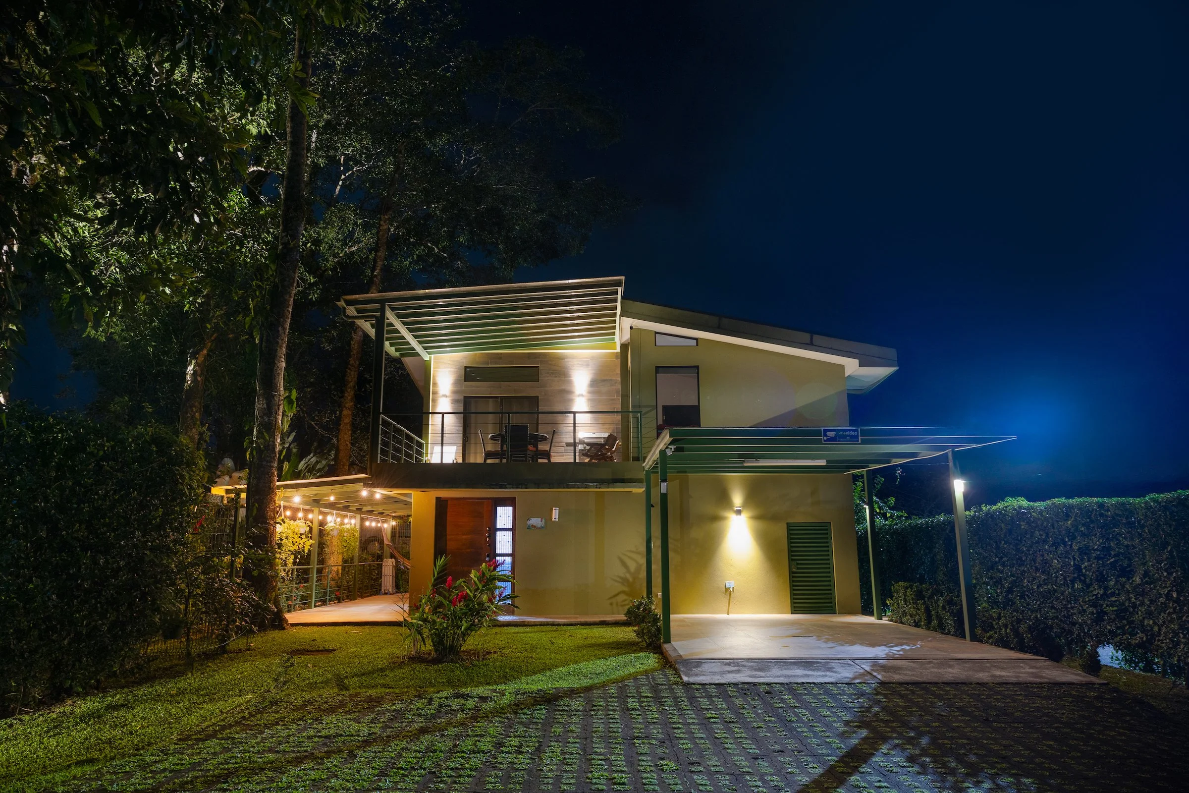 Modern two-story house at night with lit exterior lights, surrounded by green trees and plants, with patio furniture on the upper balcony.