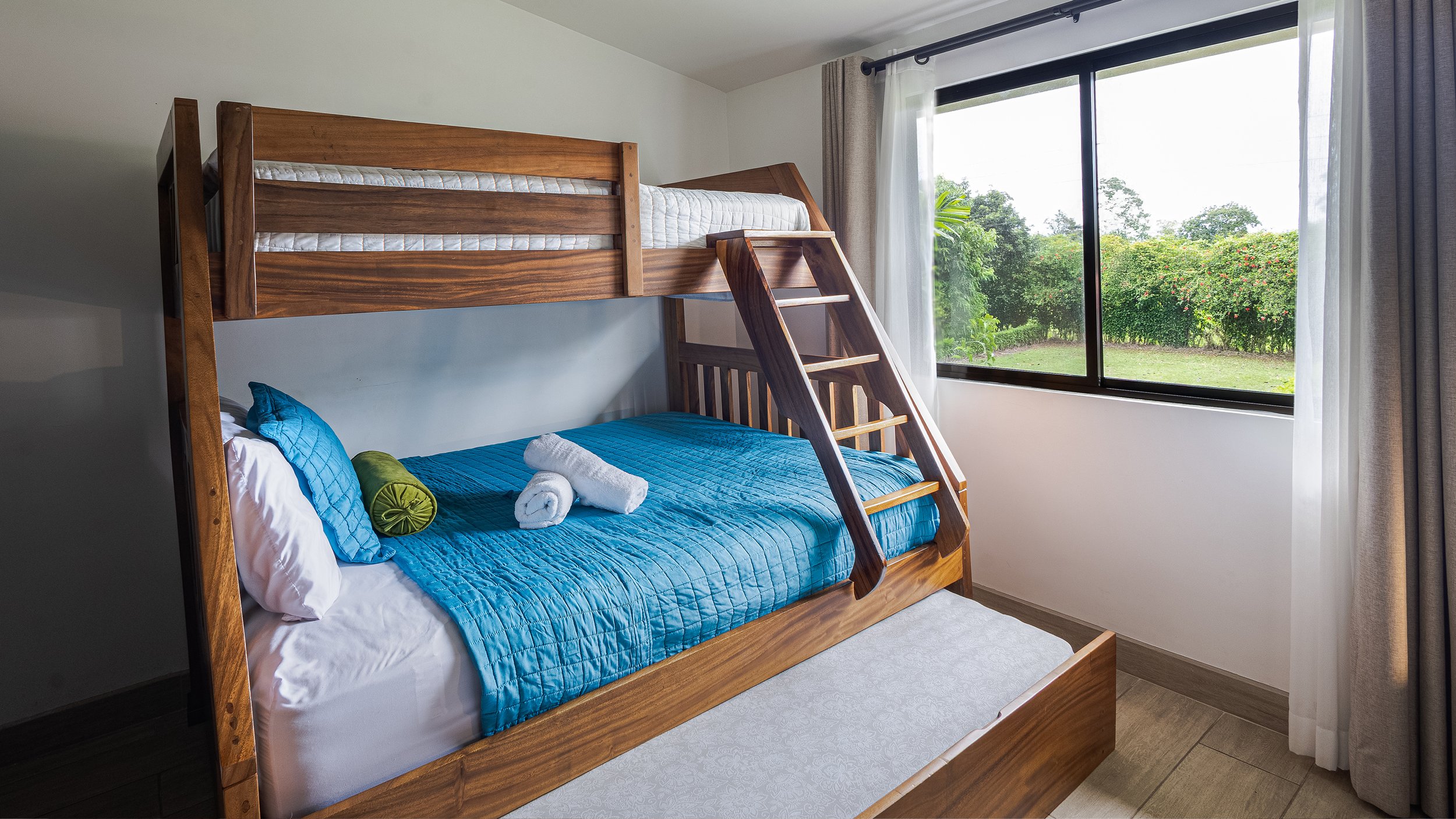 A bedroom with a wooden bunk bed next to a large window, showing green foliage outside. The bottom bunk has a blue quilt, pillows, and rolled towels, while the top bunk has a white mattress. Curtains are partially open.