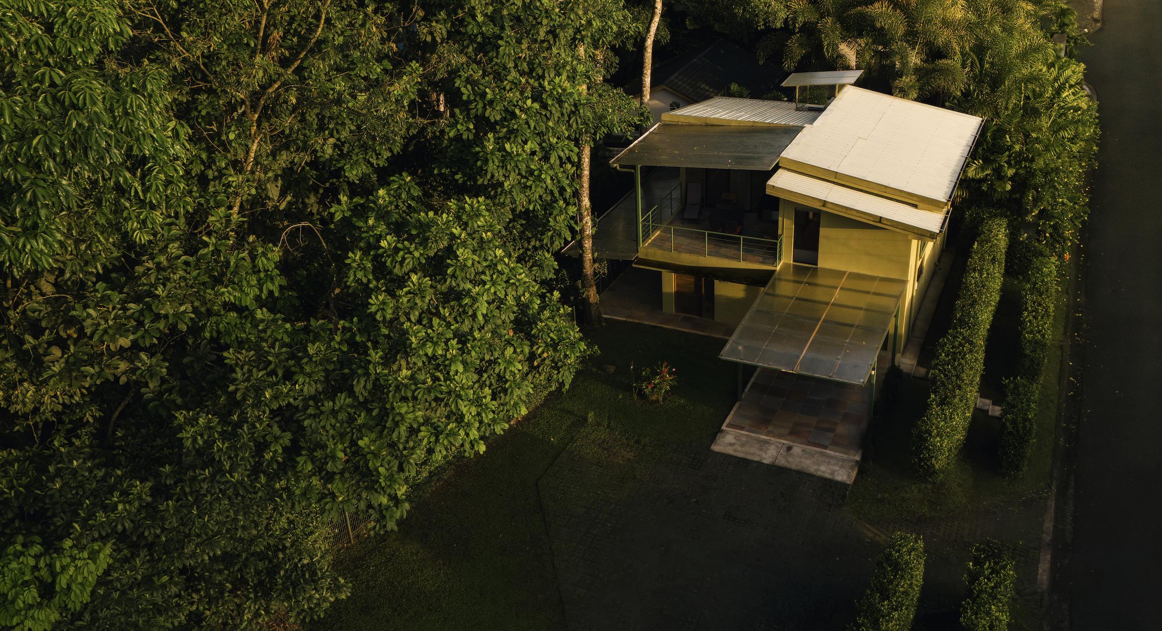 Aerial view of a modern, two-story house surrounded by lush green trees and shrubs, with a metal roof and a glass balcony, located next to a dark street.