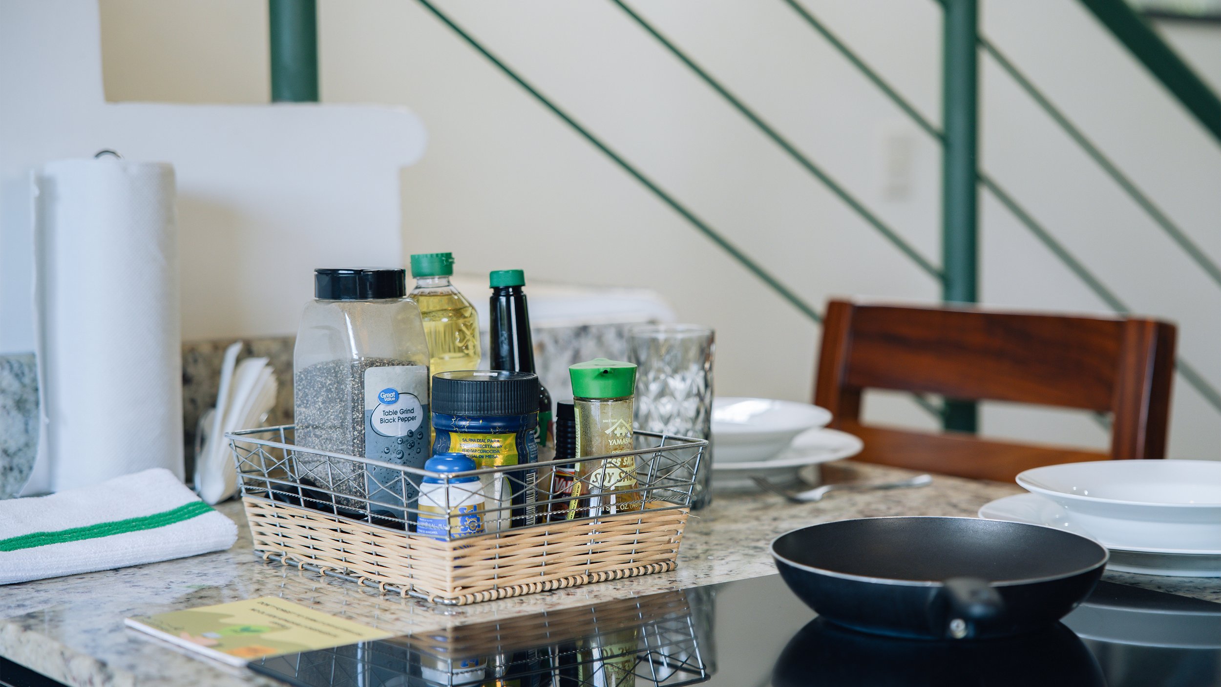 Kitchen countertop with a wire basket containing various spices, oils, and seasonings, near paper towels, plates, and bowls, with a green staircase in the background.