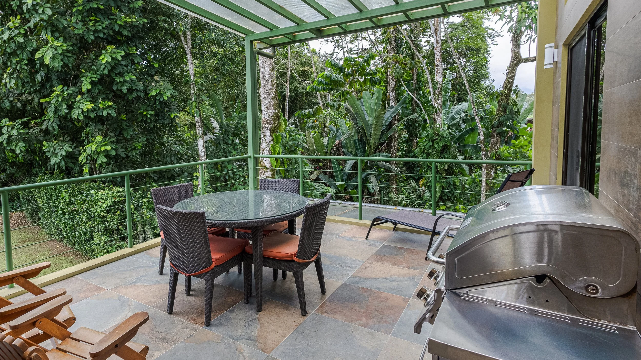 Balcony with outdoor furniture including a round glass table with four wicker chairs with orange cushions, a lounge chair, a metal grill, and lush green trees in the background.