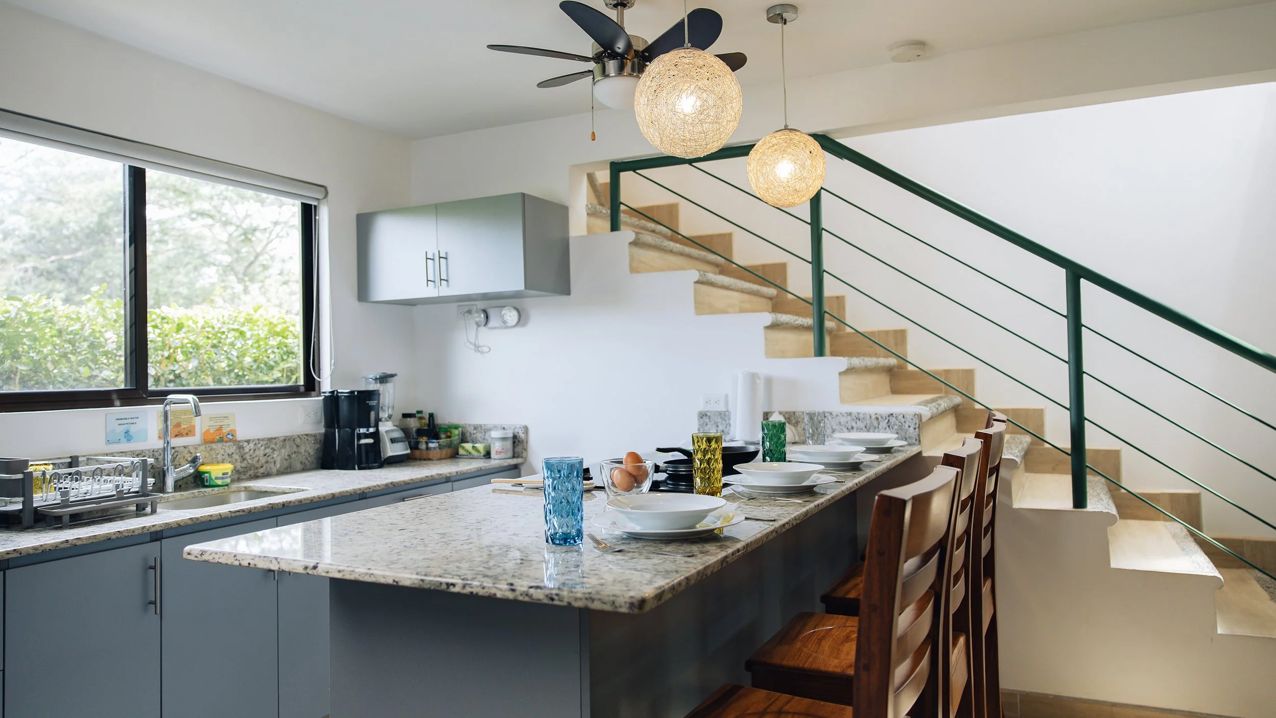 A kitchen with granite countertops, a window with a view of green foliage, and a dining area set with plates, glasses, and utensils. There are pendant lights hanging from the ceiling, a staircase with a green railing, and a ceiling fan.