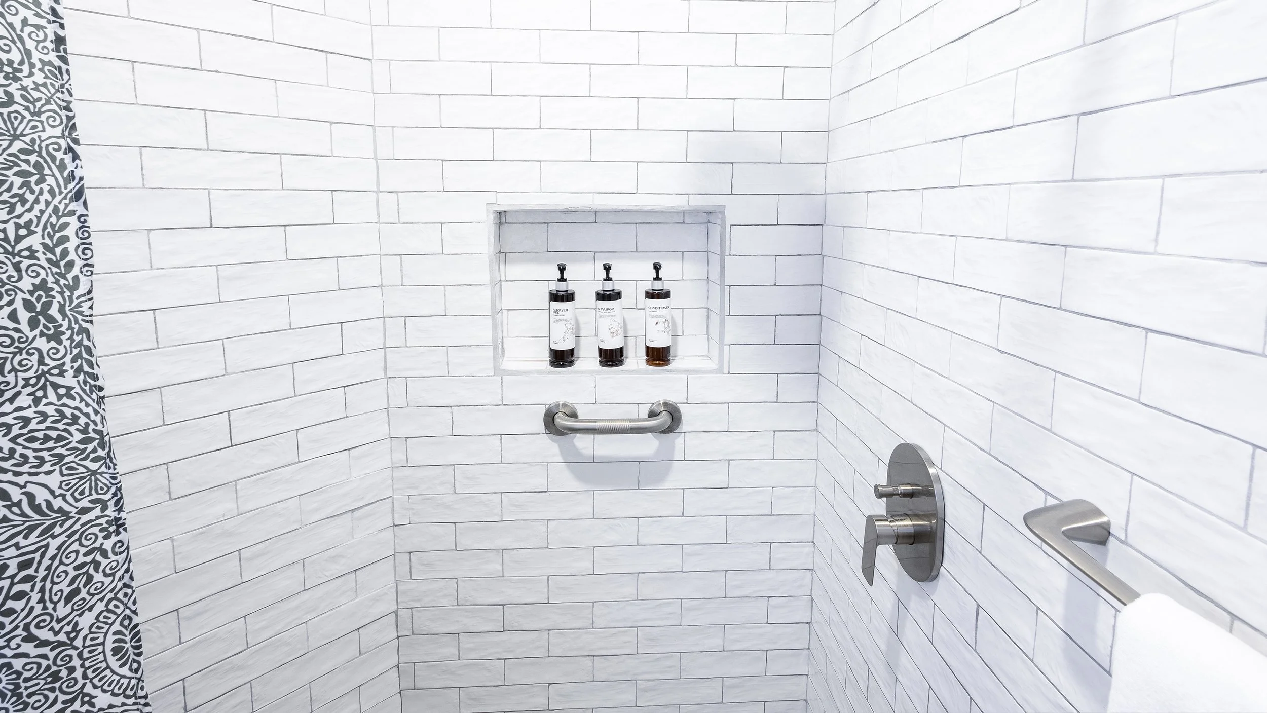 A shower with white subway tile walls, a built-in niche holding three bottles of bath products, a metal grab bar, and a shower faucet.