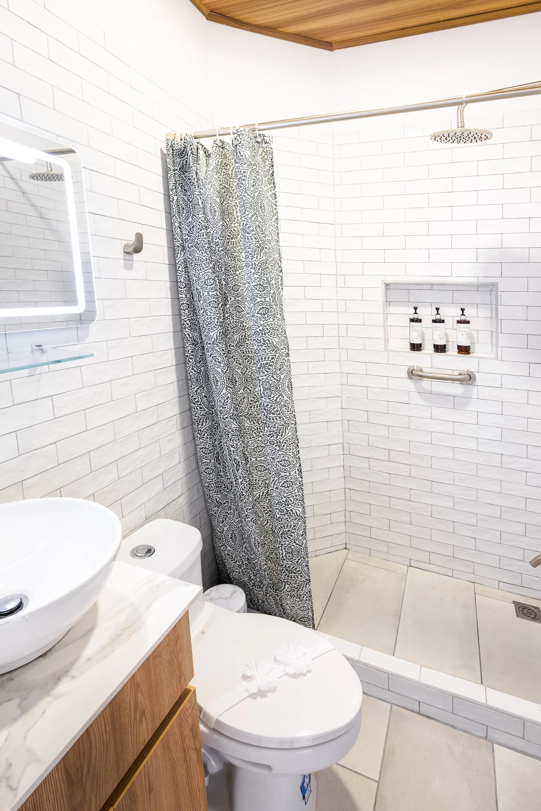 A clean bathroom featuring a toilet with a closed lid, a wooden cabinet with a marble countertop and vessel sink, a mirror with lighting, patterned shower curtain, built-in shelf with toiletries, and tiled shower area with a showerhead and grab bar.