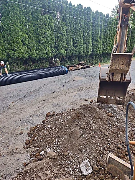 Construction site with an excavator, dirt pile, black corrugated pipe, and green trees in the background.
