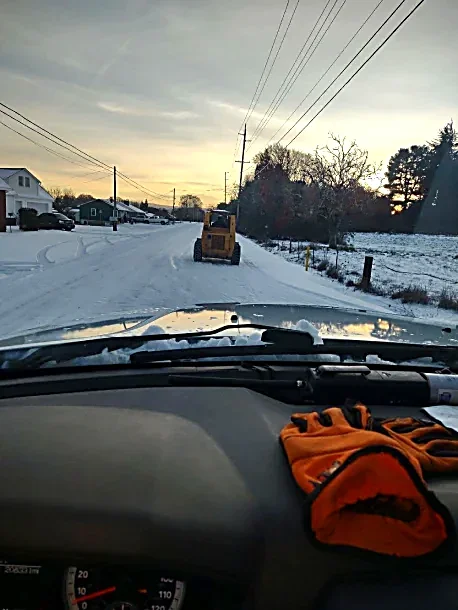 View from inside a vehicle driving on a snowy residential street during sunset with a road construction vehicle ahead, and orange gloves on the dashboard.