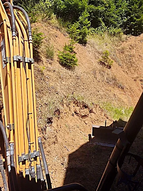 View from inside a construction vehicle showing hydraulic parts on the left and a dirt hillside with small trees on the right.