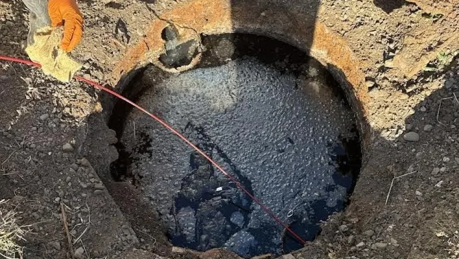 A worker's hand wearing an orange glove holding a high-pressure hose directed at a large dug hole filled with dark water and mud.