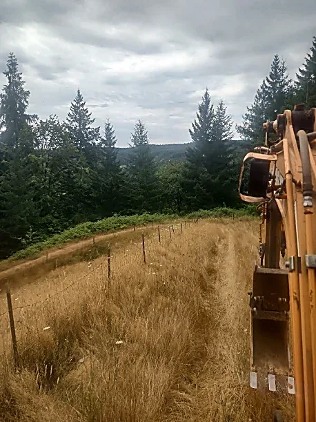 View of a dirt path through a grassy field with a barbed wire fence, surrounded by forested trees under cloudy sky, with part of construction equipment visible on the right side.