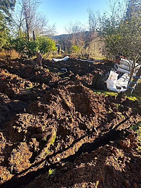 A yard with disturbed soil and construction debris, including a sofa, bags, and a ladder, with trees and hills in the background.