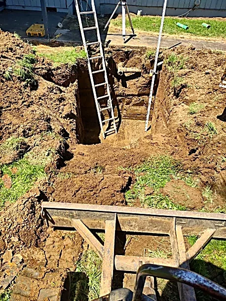 A freshly dug rectangular hole in the ground with a ladder leaning against the side and a level tool resting on top, indicating active excavation or construction work.