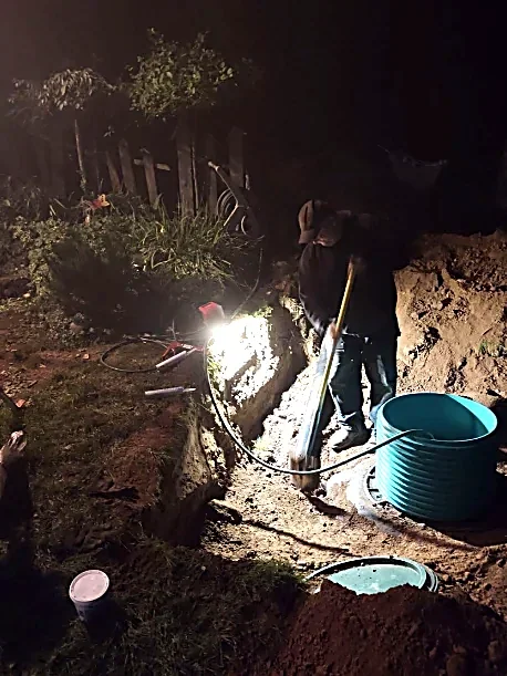 A person working on installing or repairing a pipe underground at night, using a shovel, with a large blue bucket nearby, illuminated by a flashlight or work light.