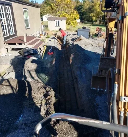 Construction site with a trench dug in the yard, a worker in a red shirt inside the trench, and construction equipment and materials around.