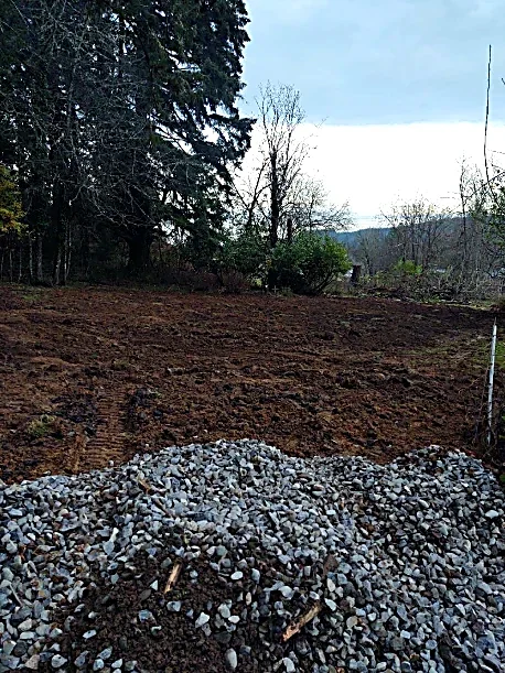 A pile of gravel in a rural outdoor area with bare trees, a patch of freshly tilled soil, and distant hills under an overcast sky.