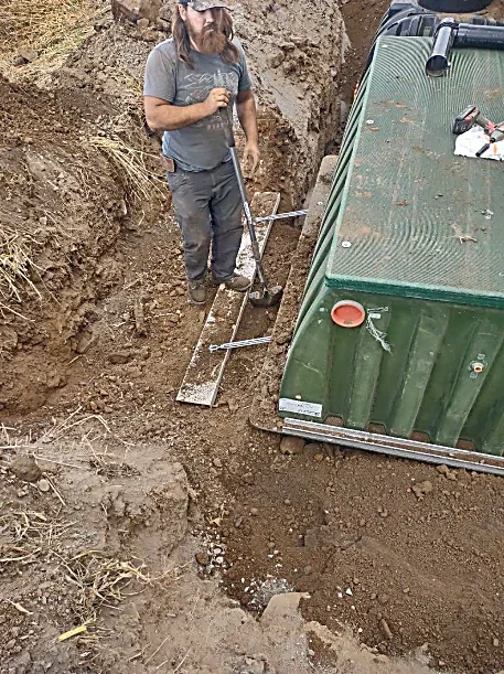 A man with long hair and a beard, wearing a baseball cap, gray t-shirt, and black pants, standing in a dirt trench holding a shovel. Next to him is a green utility vehicle or trailer.