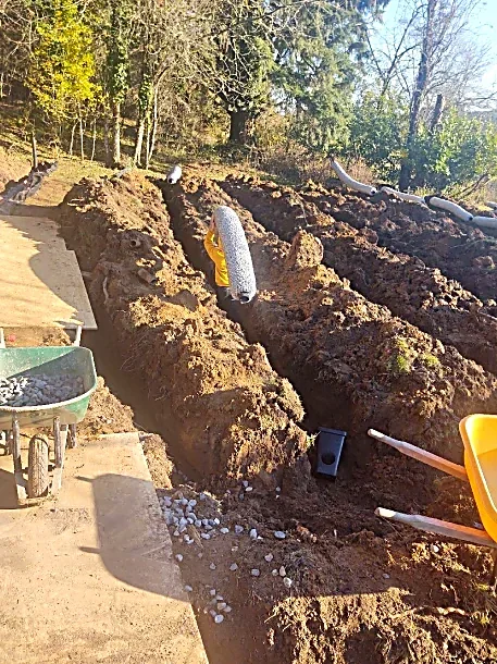 Excavation site with trenches, construction tools, a wheelbarrow, and a worker in a yellow jacket installing a large gray pipe.