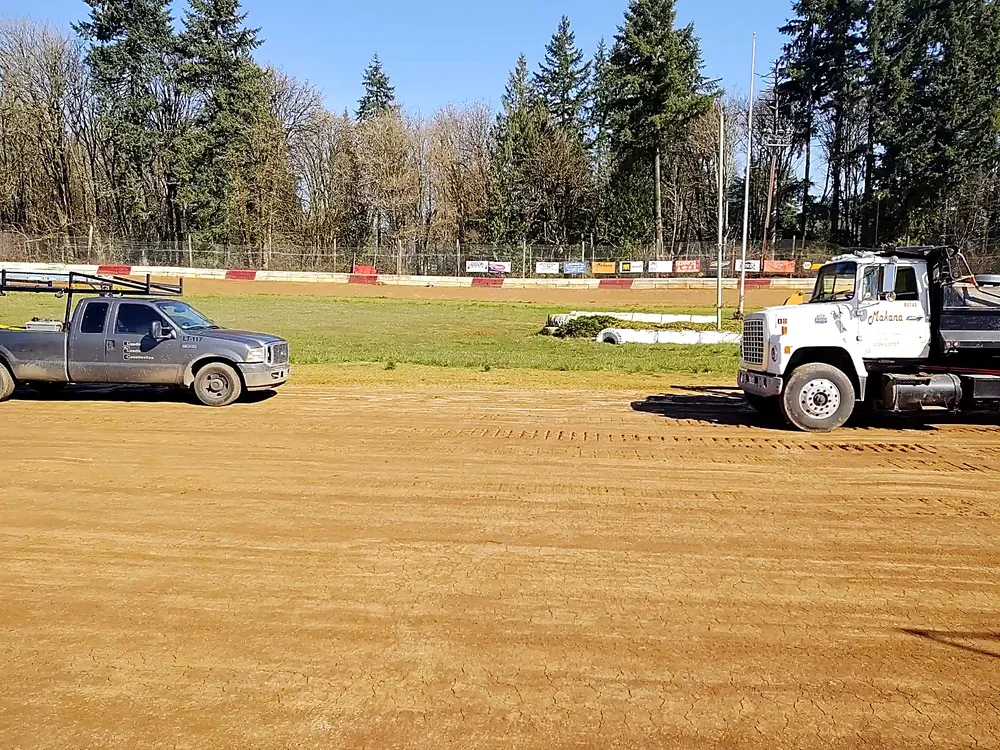 A dirt racetrack with two pickup trucks parked on the side, one gray and one white, with a wooded area and a fence in the background.