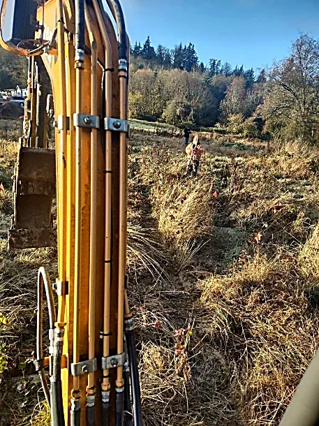 A yellow construction excavator digger with hydraulic arms in a field with shrubs and trees, and a worker in a high-vis vest in the background.