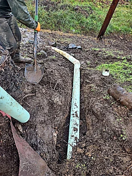 Person digging a trench for pipe repair in soil with a shovel, PVC pipe and underground utilities.