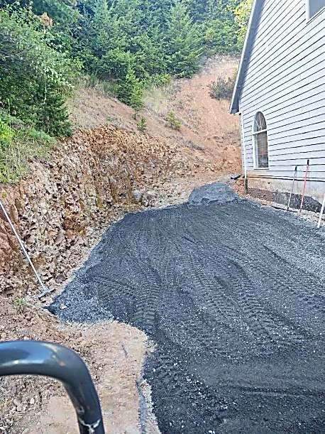 A gravel driveway under construction beside a house, with a hill and trees in the background.