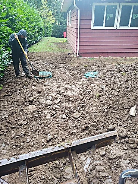 Person using a shovel to work on a cleared and excavated yard beside a red house with white windows.