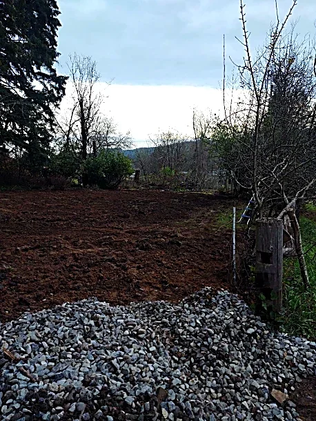 A yard with freshly turned soil, a pile of gravel in the foreground, and trees and cloudy sky in the background.