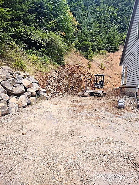 A small construction site with a mini excavator and piles of rocks on the lot, next to a house, surrounded by trees and a hillside.