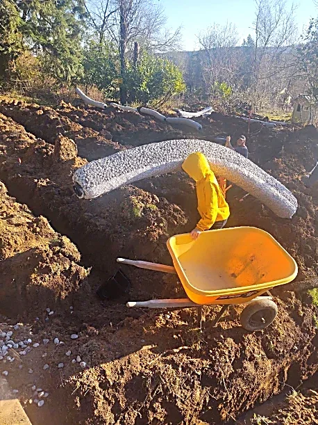 Person in yellow raincoat moving soil with a yellow wheelbarrow during outdoor construction or gardening work.
