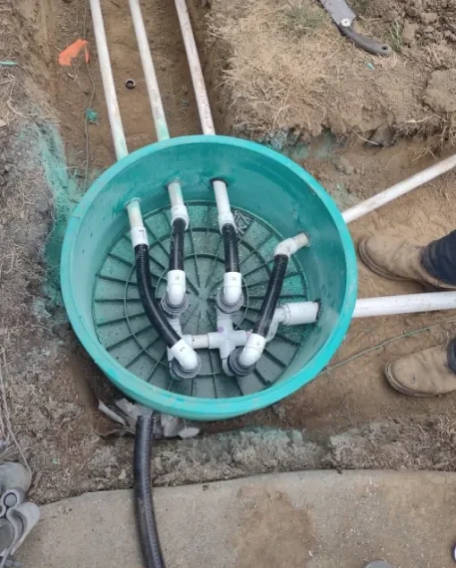 A green utility box with multiple pipe connections and black surge protectors inside, surrounded by soil and construction tools.