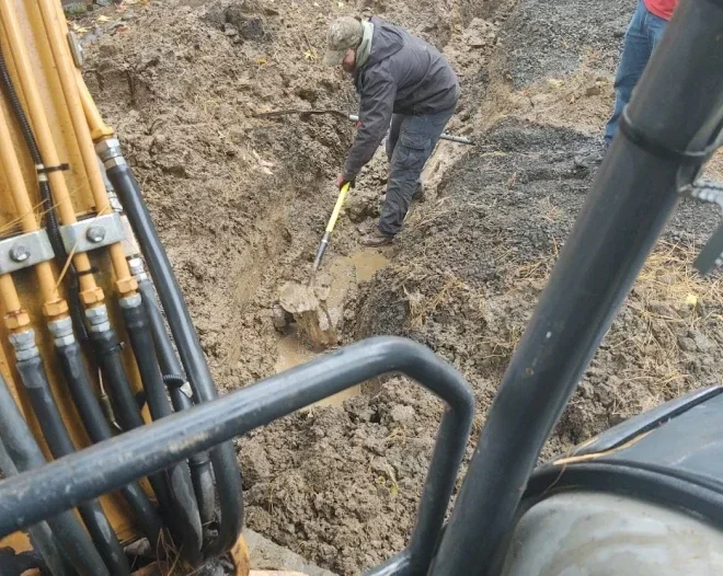 Worker in a muddy excavation site uses a shovel near a large underground pipe, with construction equipment in the foreground.