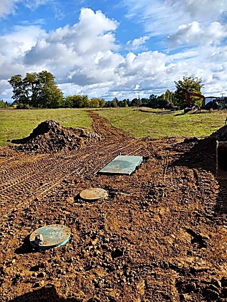 Construction site with earthwork and utility access points in a field under a cloudy sky.