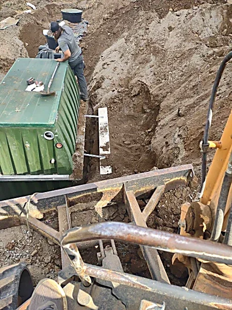 A person working on a green machine, possibly a generator or fuel tank, at a construction site with dirt and heavy machinery, including an excavator in the foreground.