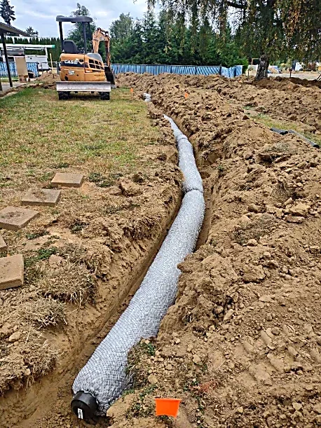 A trench with a large pipe wrapped in insulation being installed in the ground at a construction site, with a small excavator parked nearby.