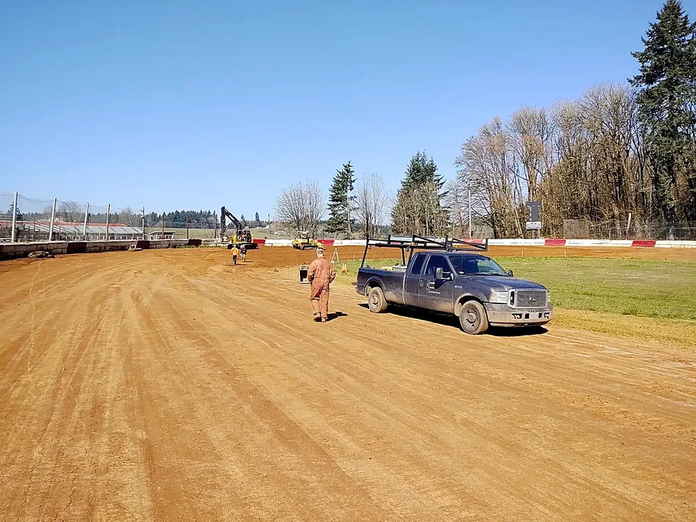 Construction workers working on a dirt racetrack with construction equipment and a pickup truck in a rural area.