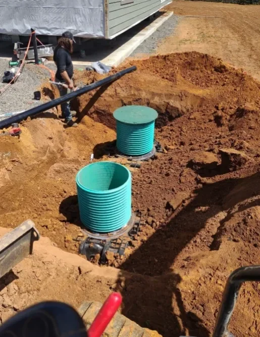 Construction site with two large green corrugated pipes installed in a dug trench, worker in dark clothing and cap standing nearby, residential building in background.