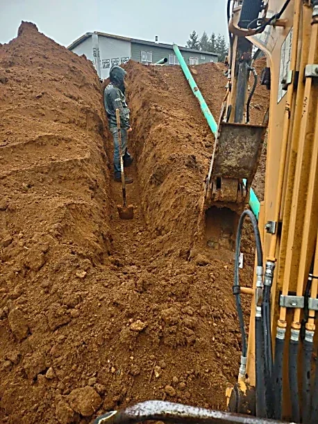A construction worker standing in a trench with an excavator nearby, surrounded by dirt and construction equipment on a construction site.