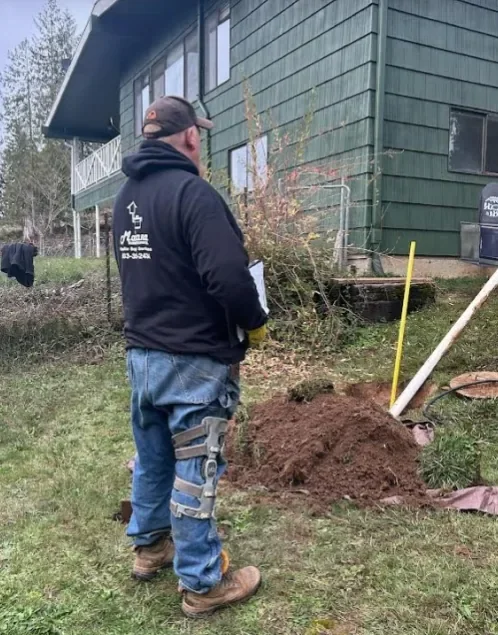 A man stands outdoors near a pile of dirt, holding a notepad, with a green house in the background.
