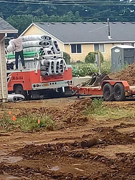 A construction site with a truck carrying large rolls of material, a person standing on the truck, and construction equipment on a dirt area near a yellow house.