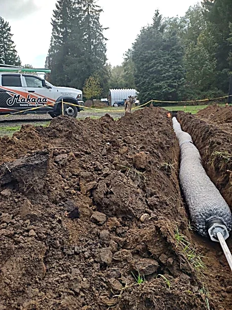 A large trench with a pipe installed inside, dirt piled on both sides, and a truck in the background with "Alakana" branding, trees, and a cloudy sky.