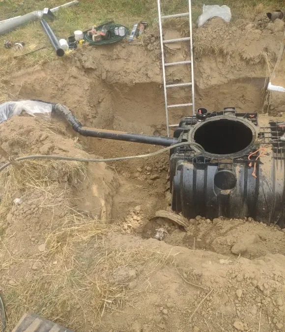A large underground septic tank with its top opening visible, partially buried in a dirt excavation site. A ladder leans against the edge of the excavation, and tools and pipes are scattered nearby.