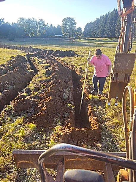 A person digging a trench in a field with a shovel next to construction equipment.