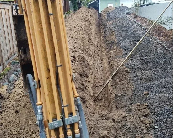 Excavation site with a trench dug in the ground for underground utilities, with a pile of dirt on each side and construction equipment nearby.