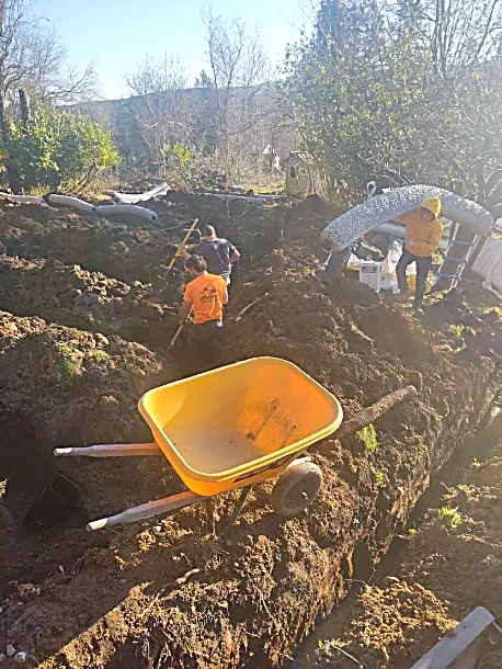 People working on a landscaping or construction project outdoors, with dirt, tools, a wheelbarrow, and trees in the background.