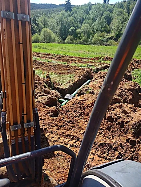 View from construction equipment showing dug trenches and pipes in a grassy field surrounded by trees and hills.