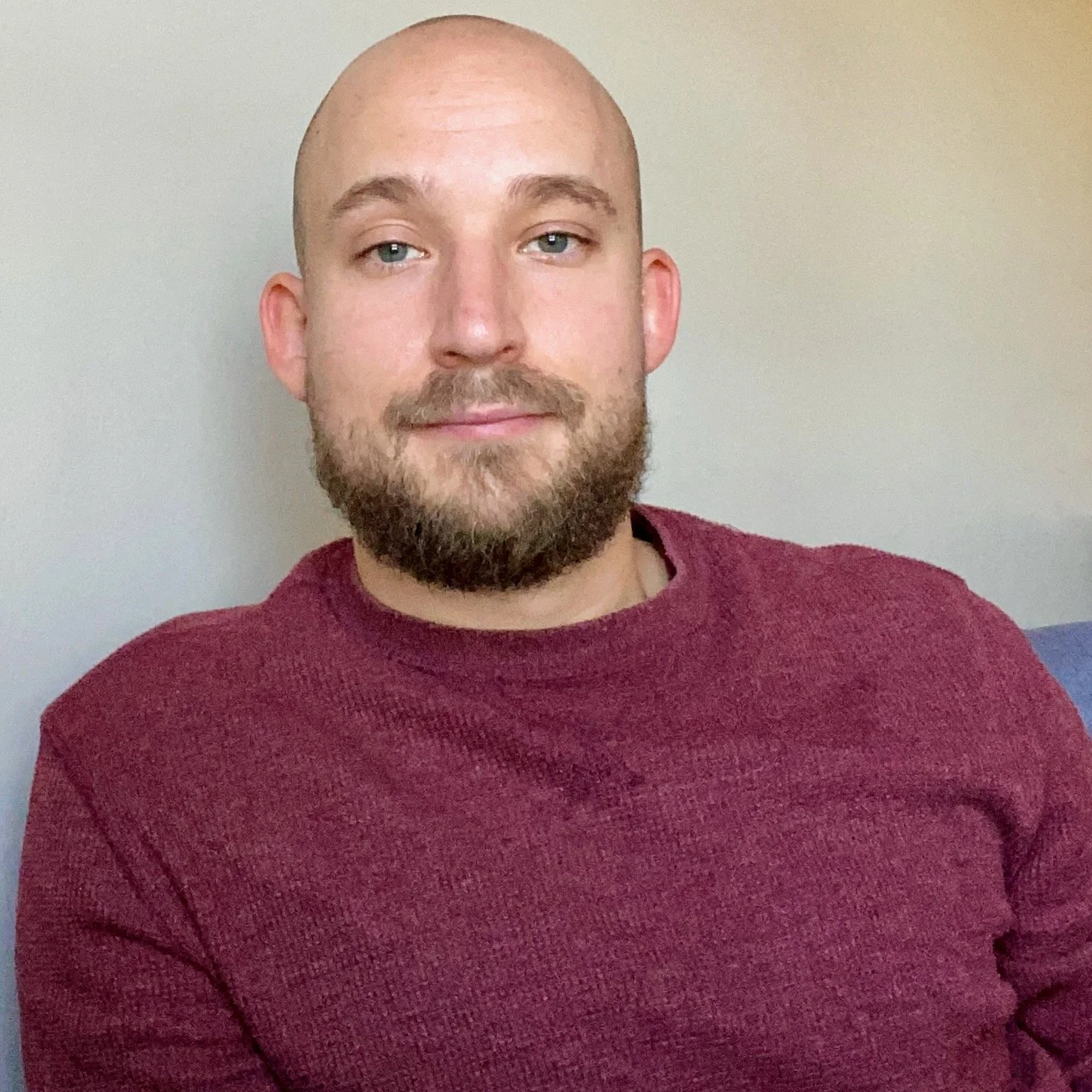A man with a shaved head and beard wearing a maroon sweater, sitting against a light-colored wall.